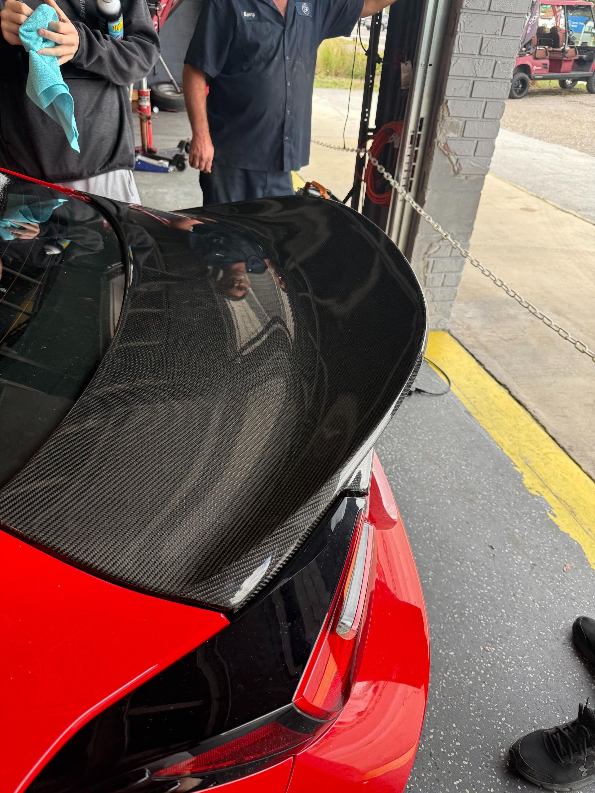 Red car with carbon fiber spoiler, inside a garage, next to a mechanic.
