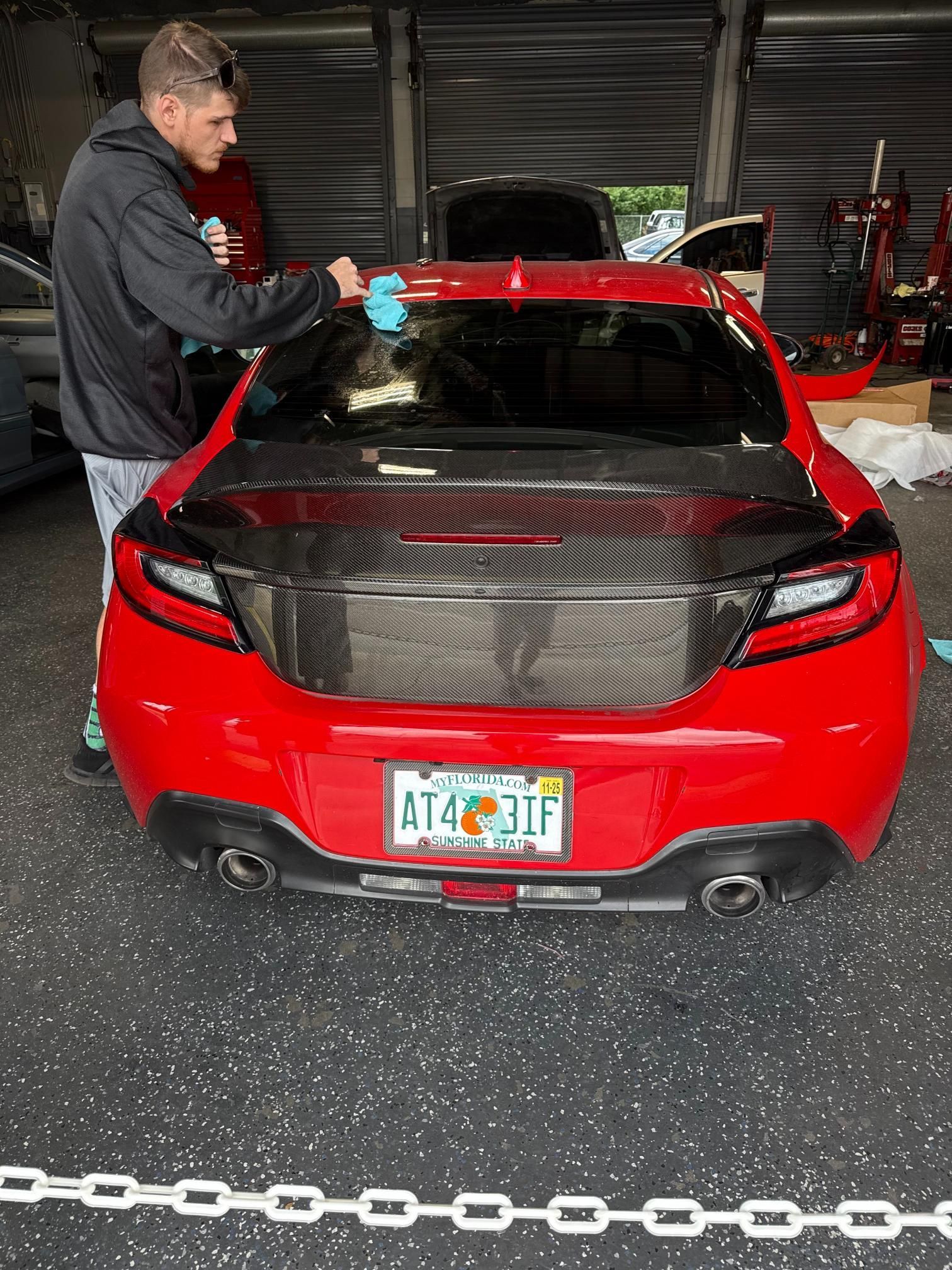 A person applying tint to a red car's rear window. The car has a carbon fiber trunk and a Florida license plate.