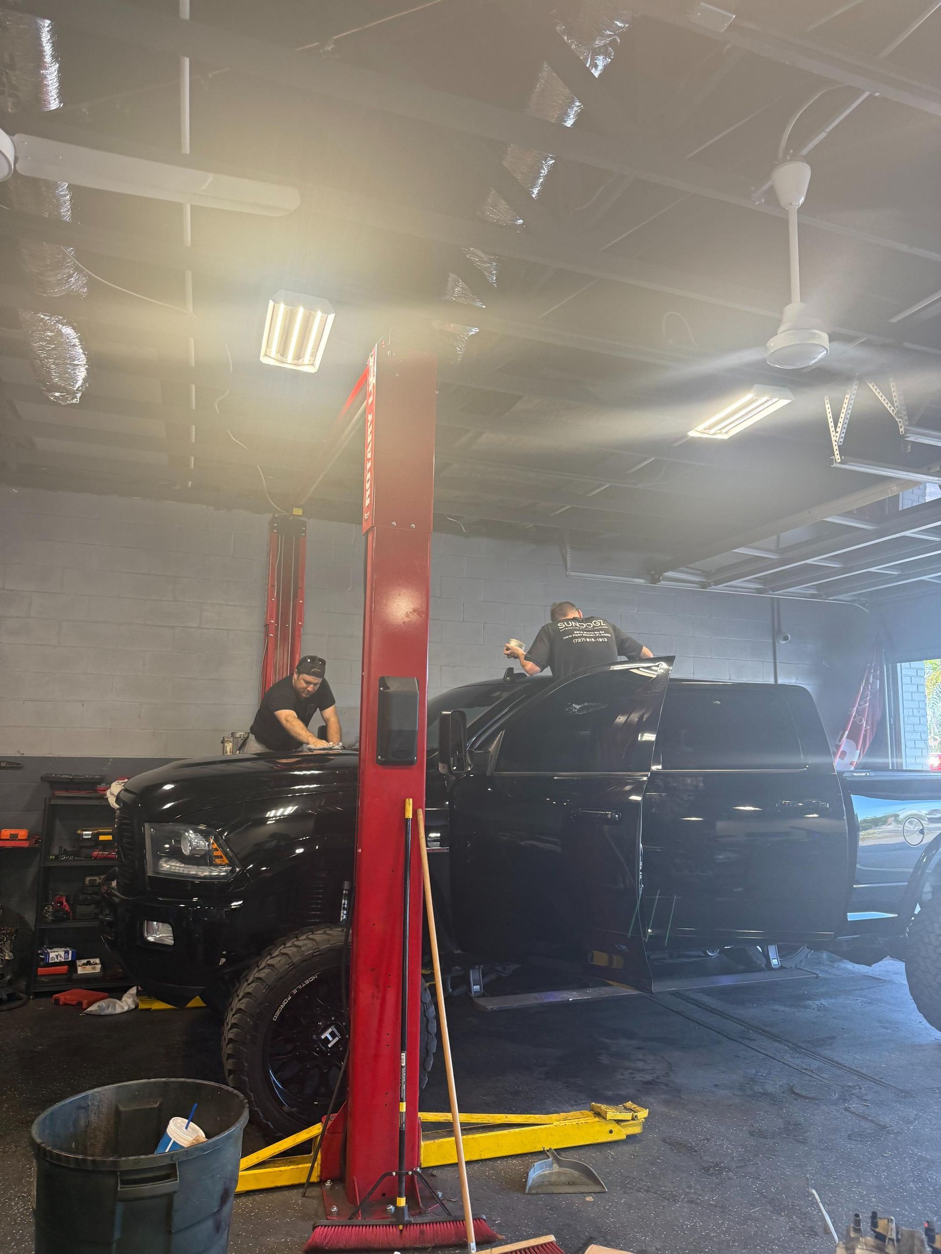 Person atop a black pickup truck in a mechanic shop, one person leans on the vehicle. Red lift posts.
