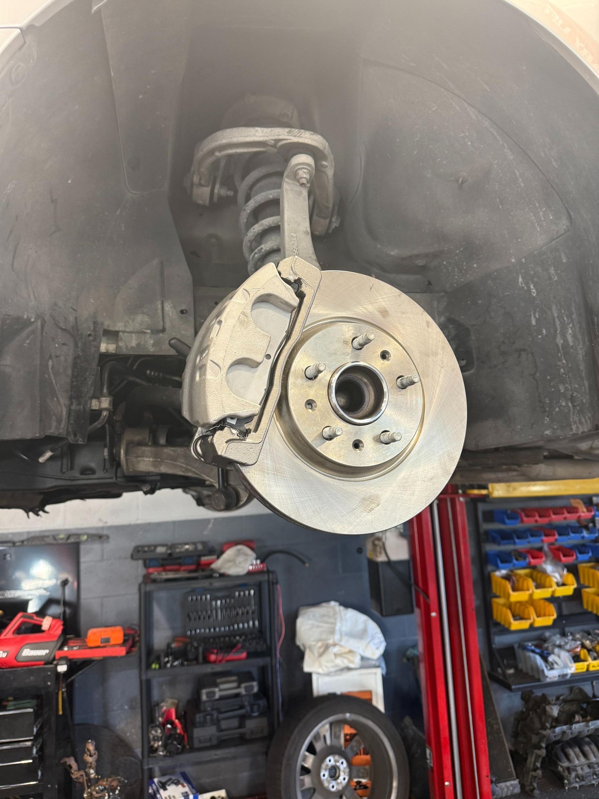 Car's front brake assembly exposed; rotor, caliper, spring strut visible; in a repair shop.