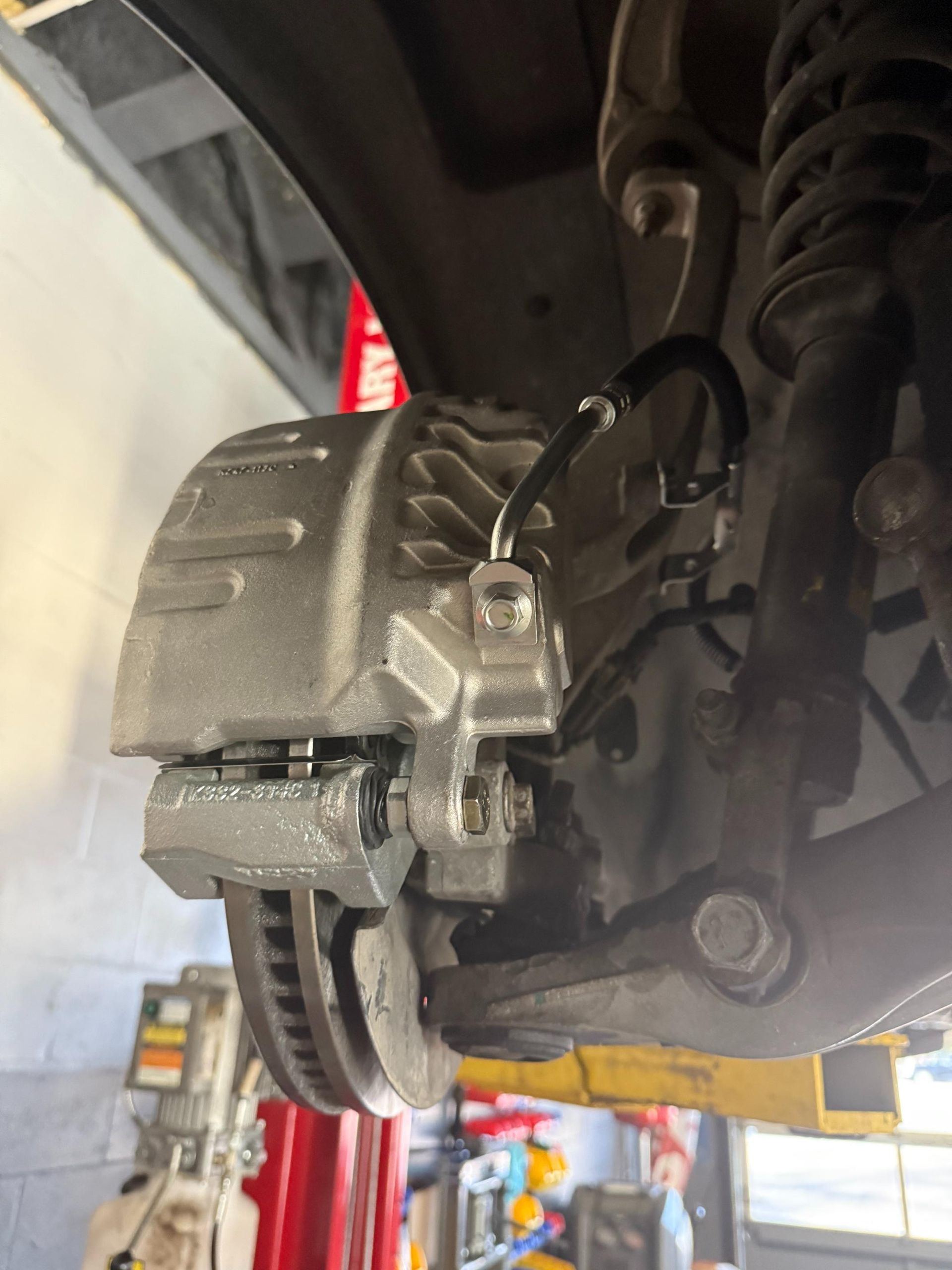 A car's front brake caliper, rotor, and suspension components are visible during service in a garage.