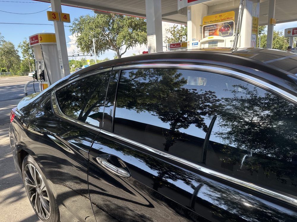 Black car parked at a Shell gas station, with tinted windows reflecting trees.