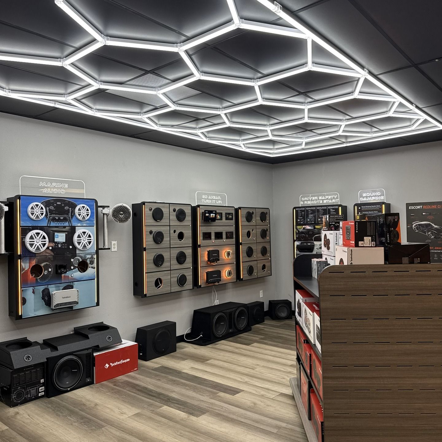 Car audio shop interior. Gray walls, speakers on display, hexagon-lit ceiling, wood flooring, and product displays.