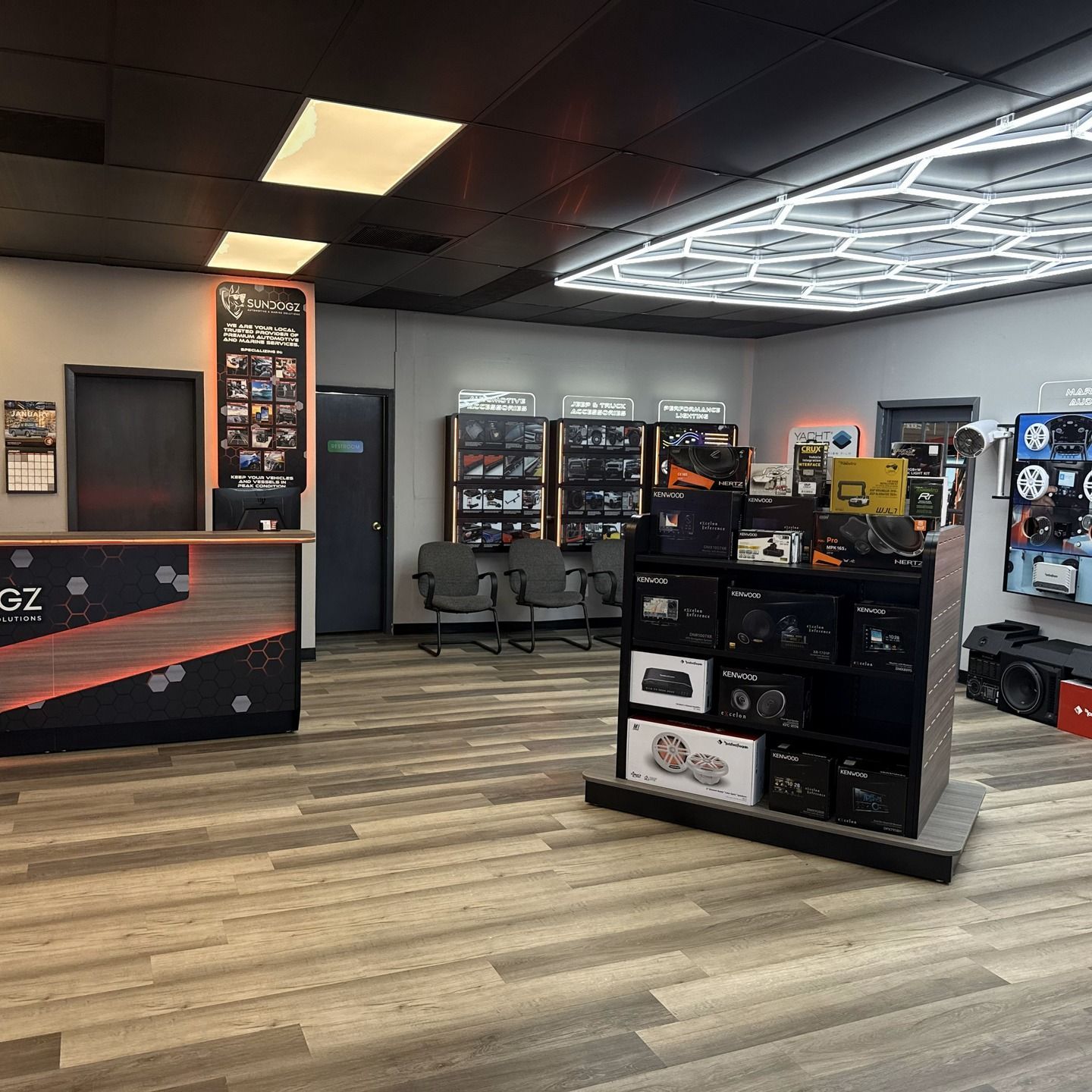 Interior of an electronics store with displays, waiting area, and counter. Wooden floor. Black and orange accents.