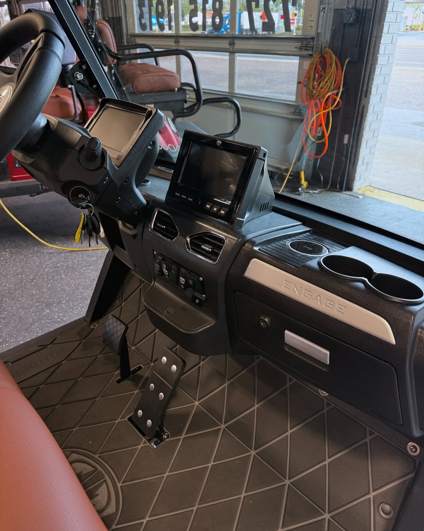 Interior of a golf cart showing a black dashboard, two screens, cup holders, and diamond-patterned floor.