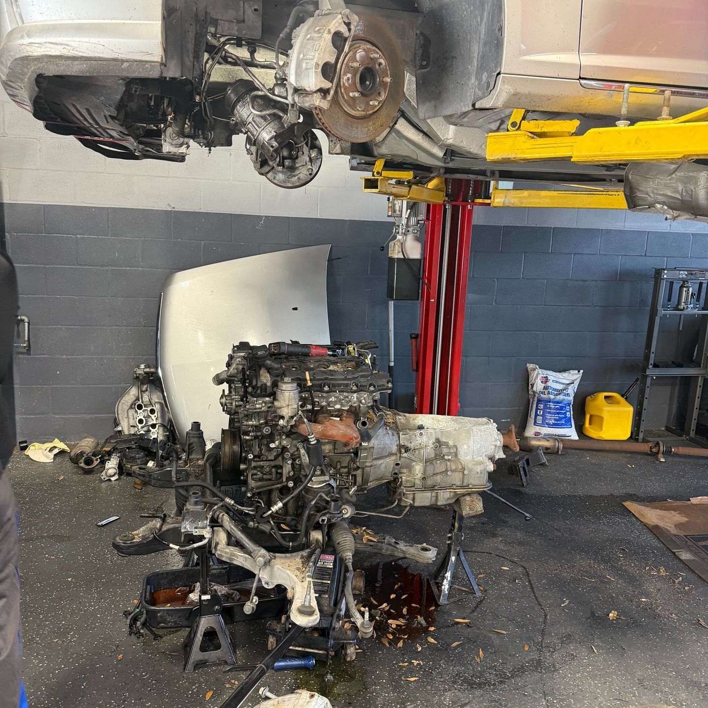 Car being repaired in a garage; engine removed and on ground. Lifted car on a hoist.