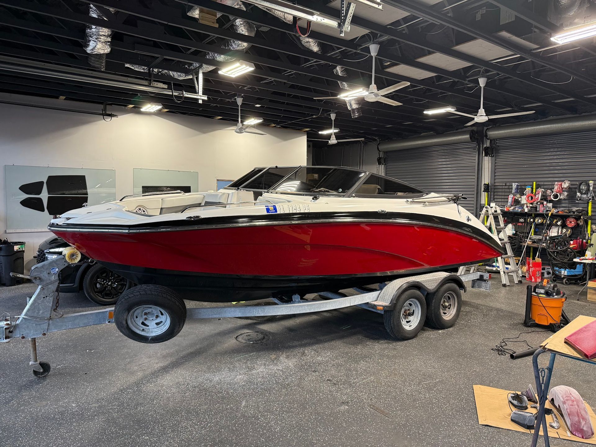 Red and white motorboat on a trailer inside a building with ceiling fans, tools and equipment visible.