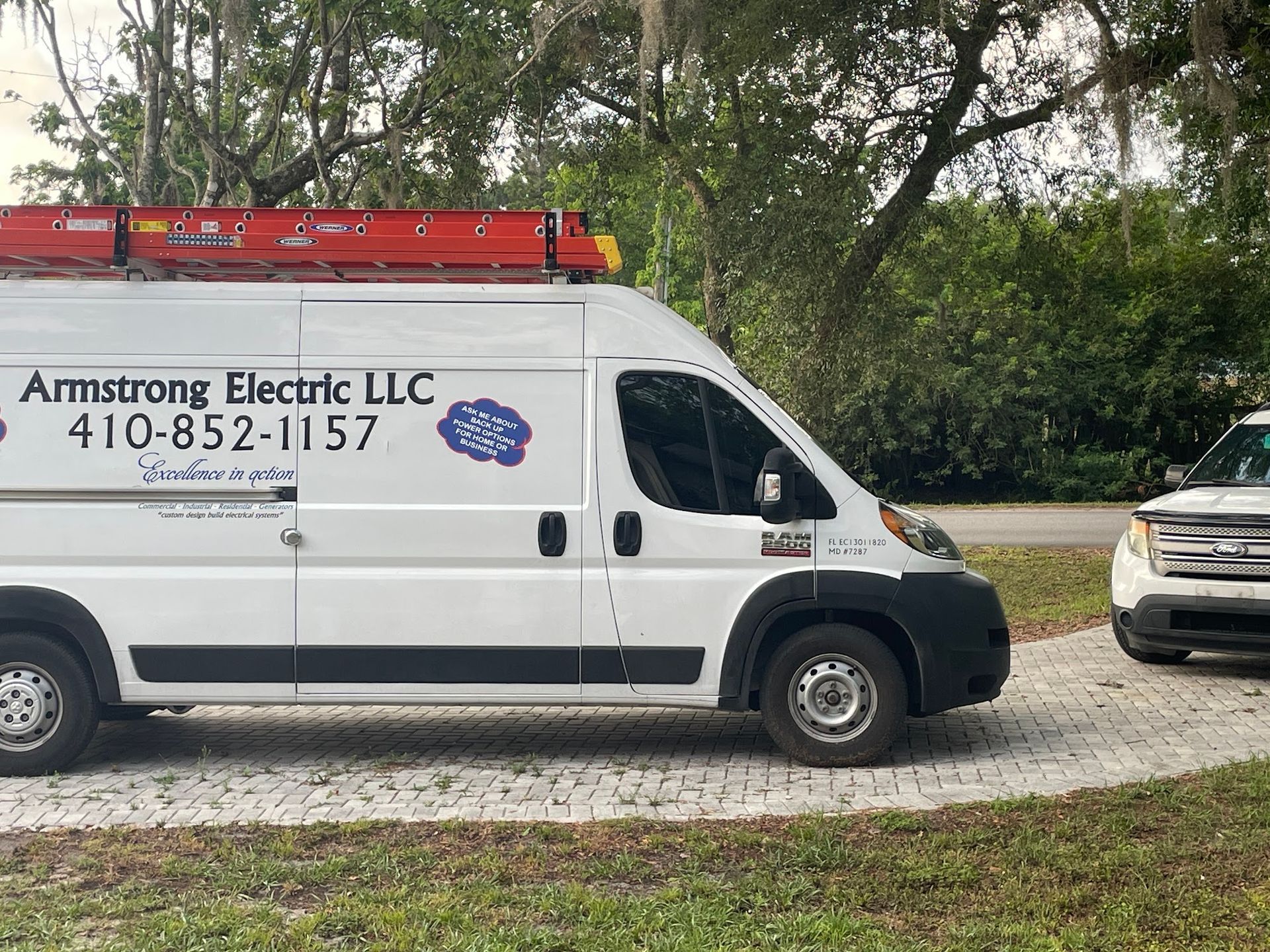White Armstrong Electric LLC van with ladder parked on a driveway next to a white SUV; green trees in the background.