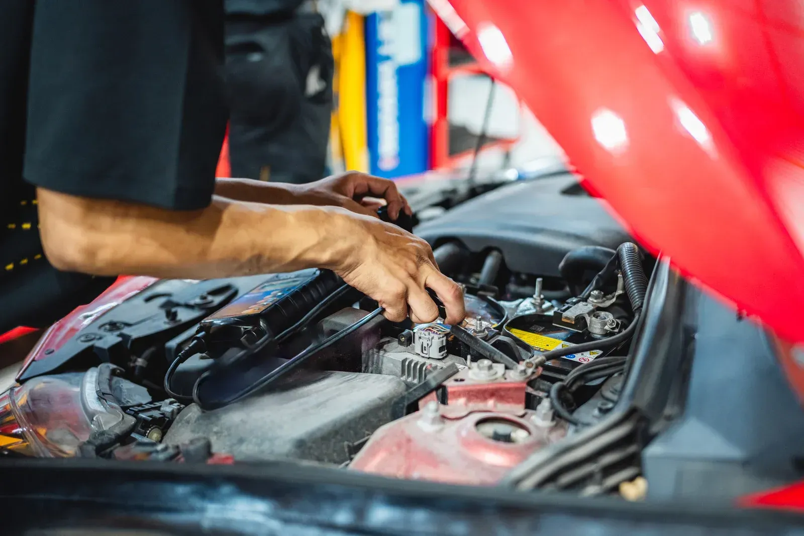 Mechanic working on a car engine under the open hood, red car, shop setting.