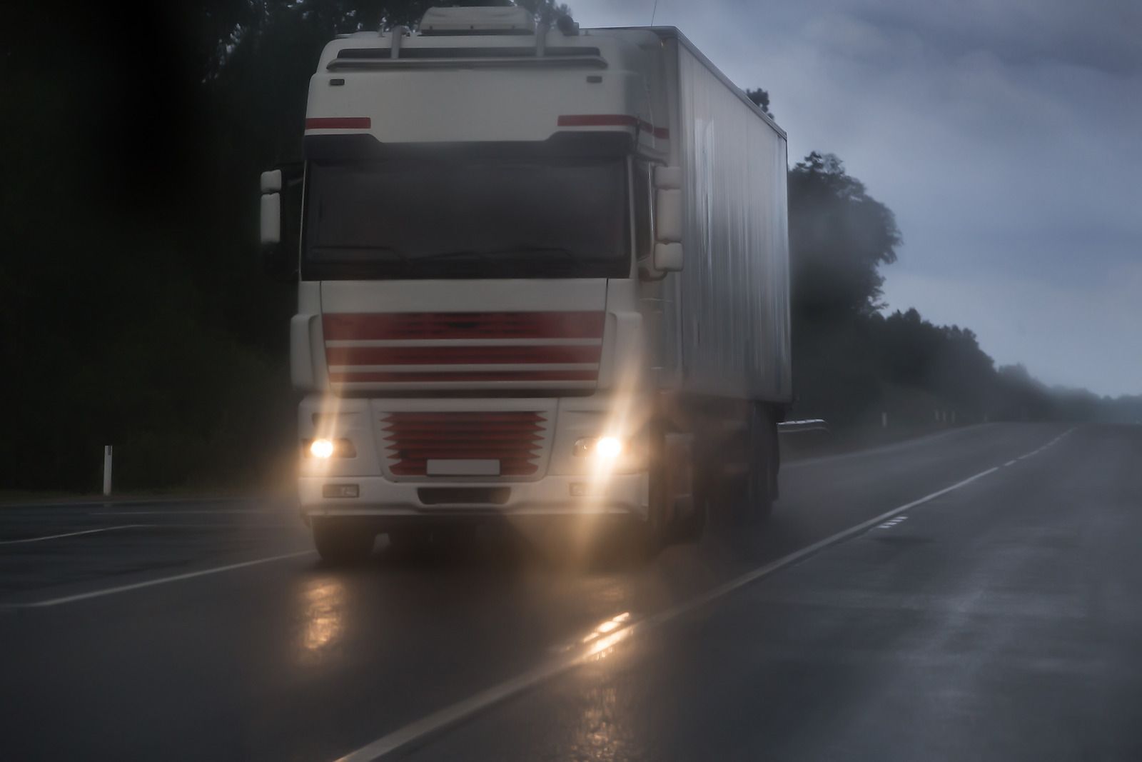 Semi-truck driving on a wet road in the rain. Headlights illuminated, dark and overcast sky.