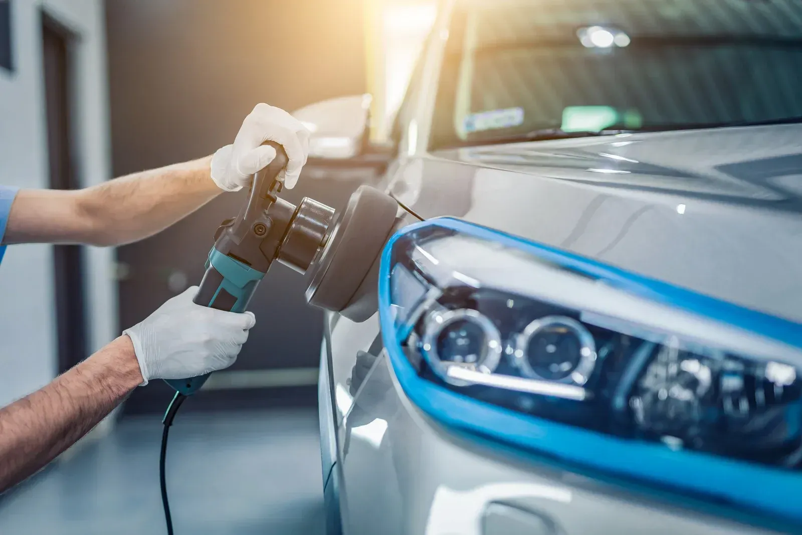 A person wearing gloves polishes a silver car's headlight with a power buffer.