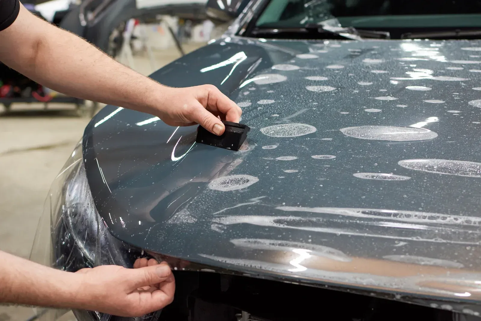 Hands applying clear film to a car hood, using a squeegee to remove air bubbles.