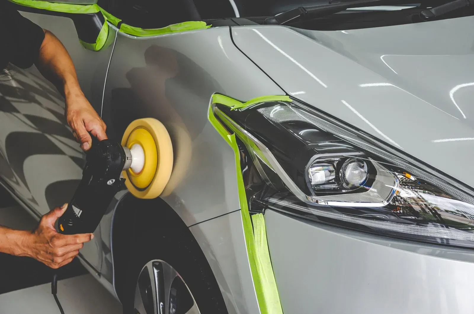 Person polishing a silver car's hood with an electric buffer, taped edges for protection, bright light.