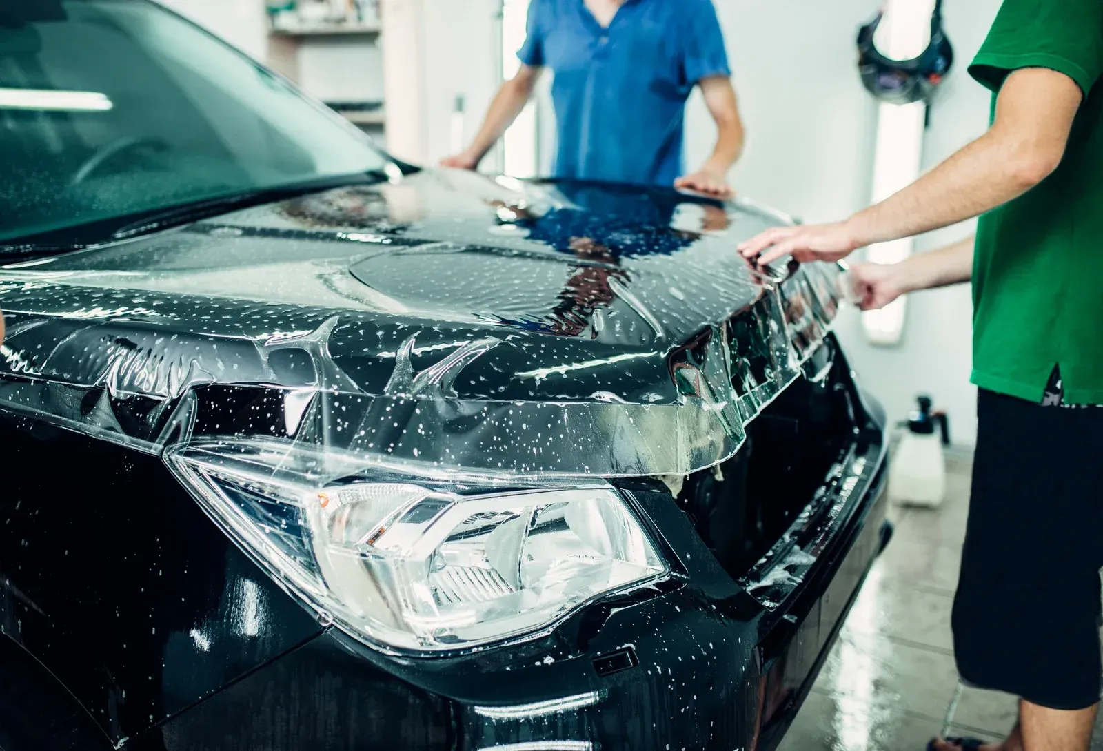 Two people applying protective film to a black car's hood in a garage.