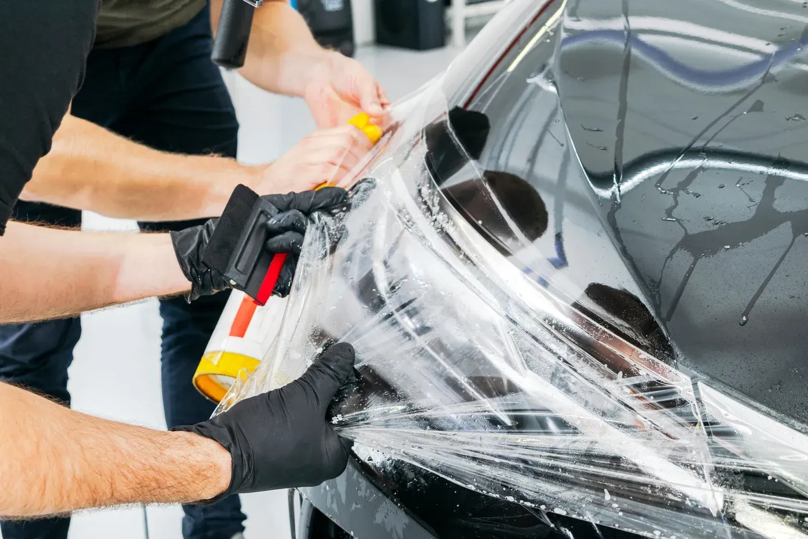 Two people applying clear protective film to a black car's fender, using tools and spray bottle.