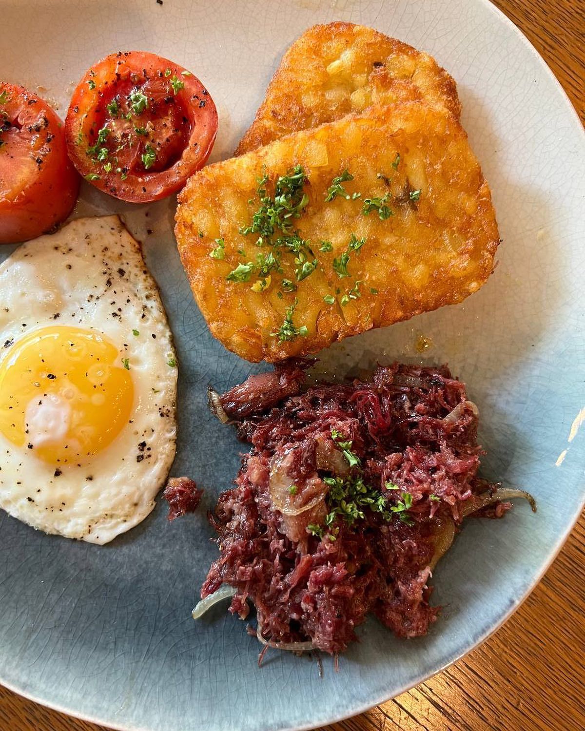 A plate of food with eggs , hash browns , tomatoes and corned beef on a wooden table.