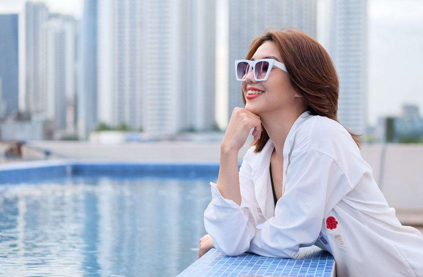 Woman in white shirt and sunglasses smiling by a blue pool with city skyscrapers in the background