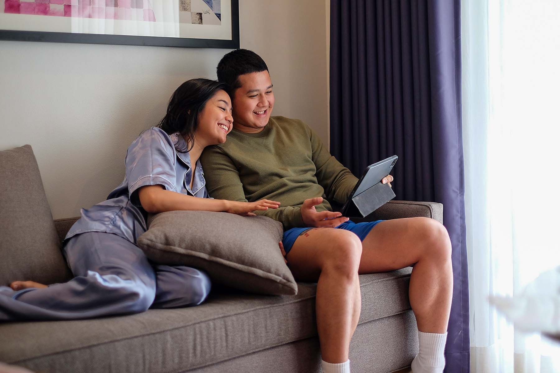 Couple on couch looking at a tablet, indoors. Woman in pajamas, man in shorts, smiling.