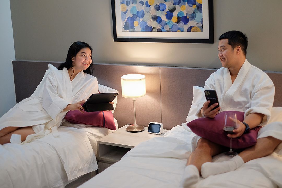 Couple in robes, in a hotel room, sitting on separate beds, using phone and tablet, smiling.
