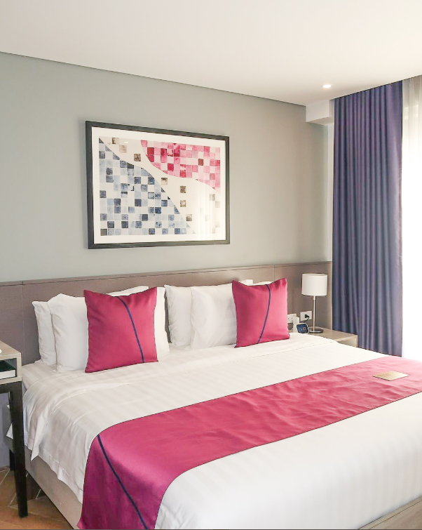 A hotel bedroom with a white bed, pink decorative pillows and runner, a framed abstract print, and purple curtains.