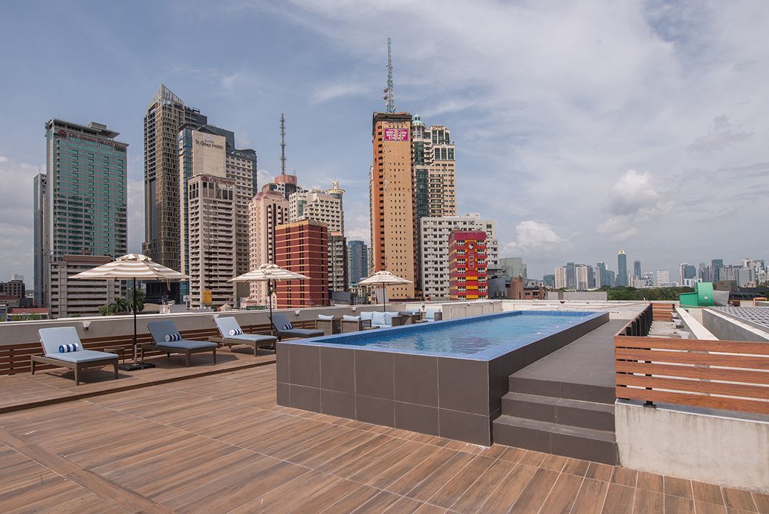 Rooftop pool with cityscape background, featuring lounge chairs, umbrellas, and wooden deck.