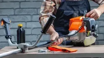 Person working on a weed wacker, holding a fuel line. Brick wall background.