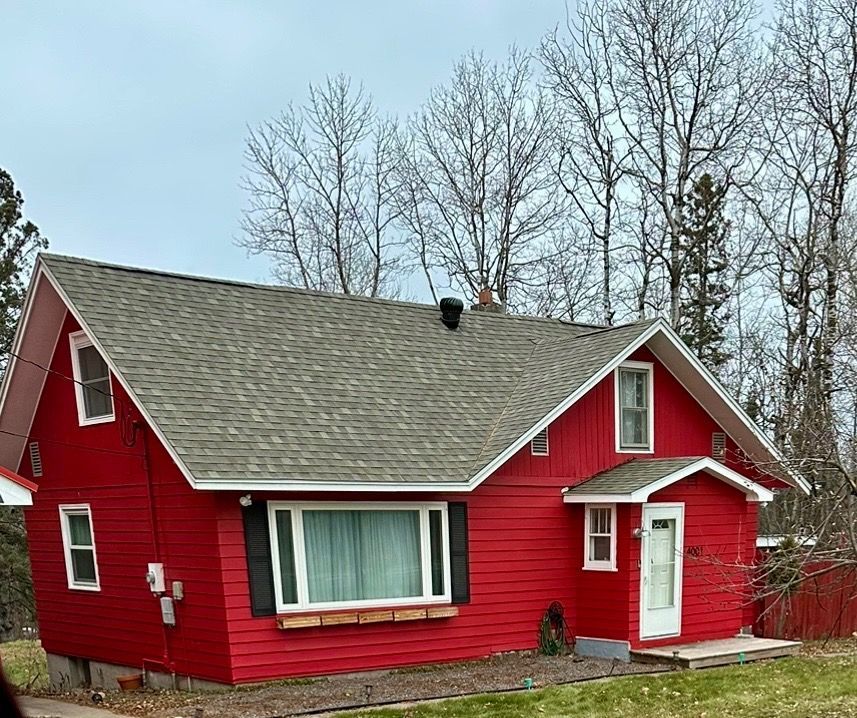 A bright red, two-story house with a grey shingled roof, set against a backdrop of bare trees under an overcast sky.