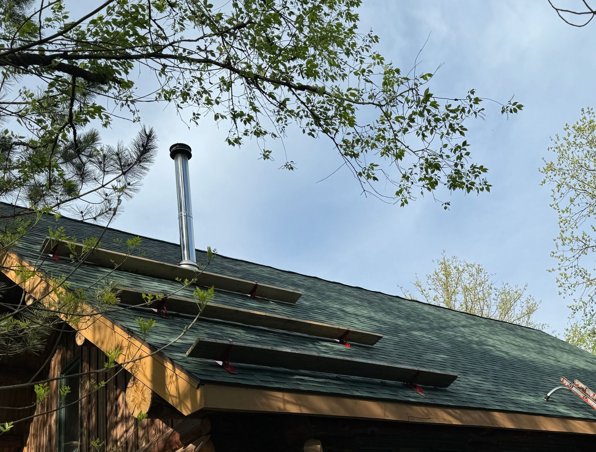 A metal chimney pipe protrudes from a green shingled roof with several horizontal mounting brackets, set against trees.