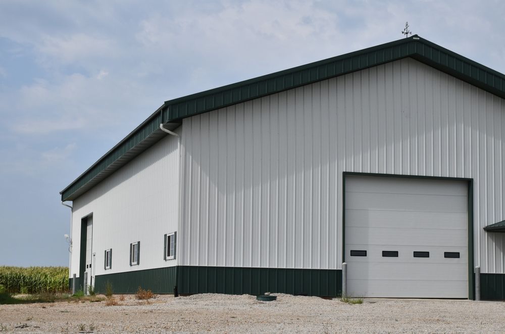 A white metal pole barn with green trim and a single large garage door, situated on a gravel lot under a blue sky.