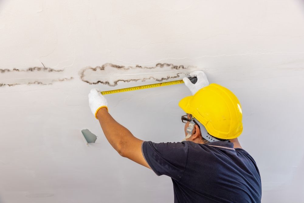 A person in a yellow hard hat and white gloves uses a measuring tape to inspect a damaged area on a white ceiling.
