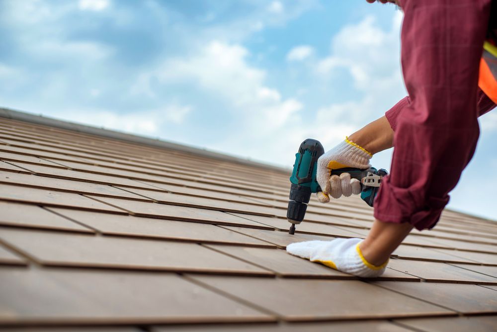A person wearing gloves uses a power drill to install brown roof tiles against a blue, cloudy sky.