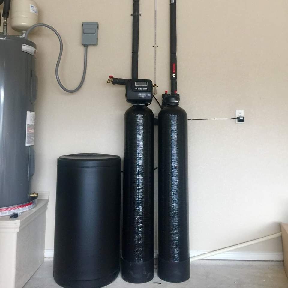 Water softener system in a garage; includes a water heater, two tall black tanks, and a round salt storage tank.