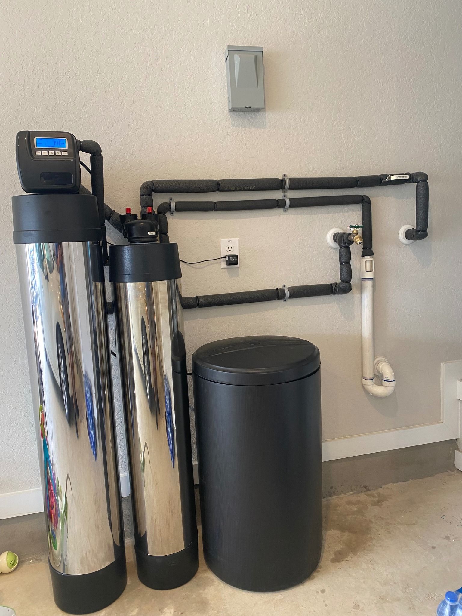 Water filtration system installed in a garage with two silver tanks, a black tank, and associated plumbing against a wall.
