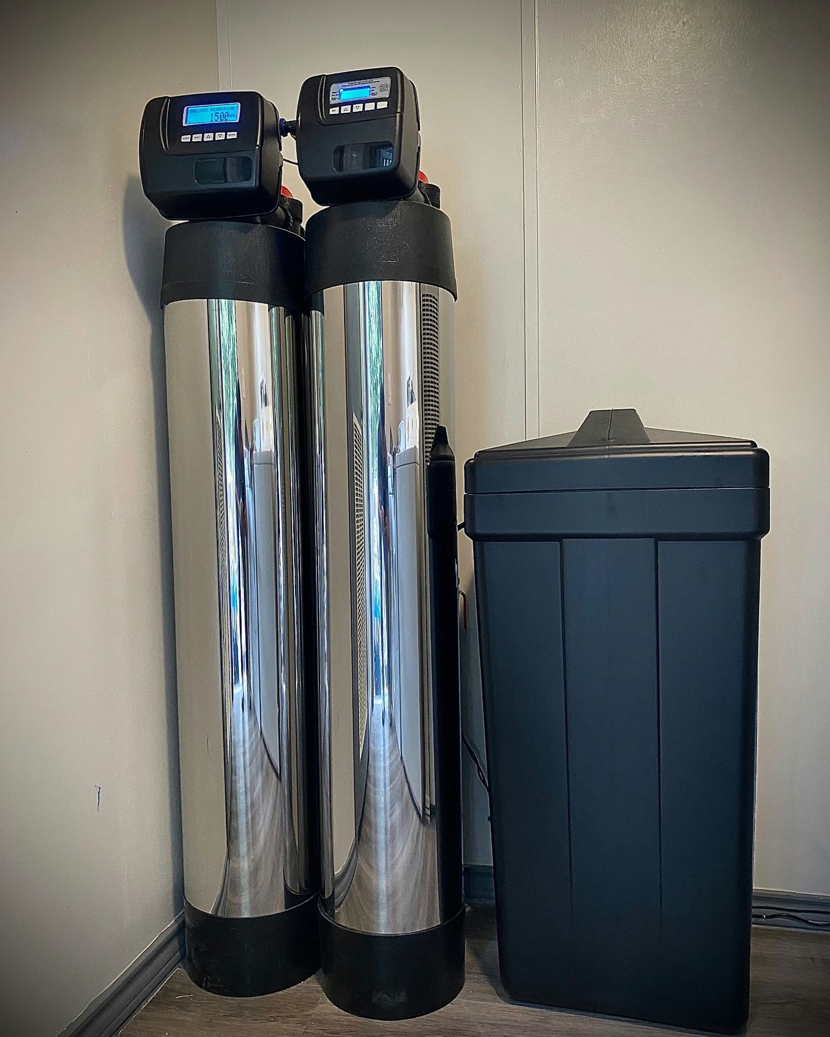 Two tall, silver water filtration tanks with displays next to a black salt storage container in a corner against