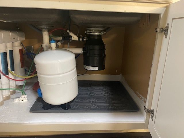 Under-sink cabinet with a garbage disposal, white water tank, and filter system. A gray mat sits on the cabinet floor.