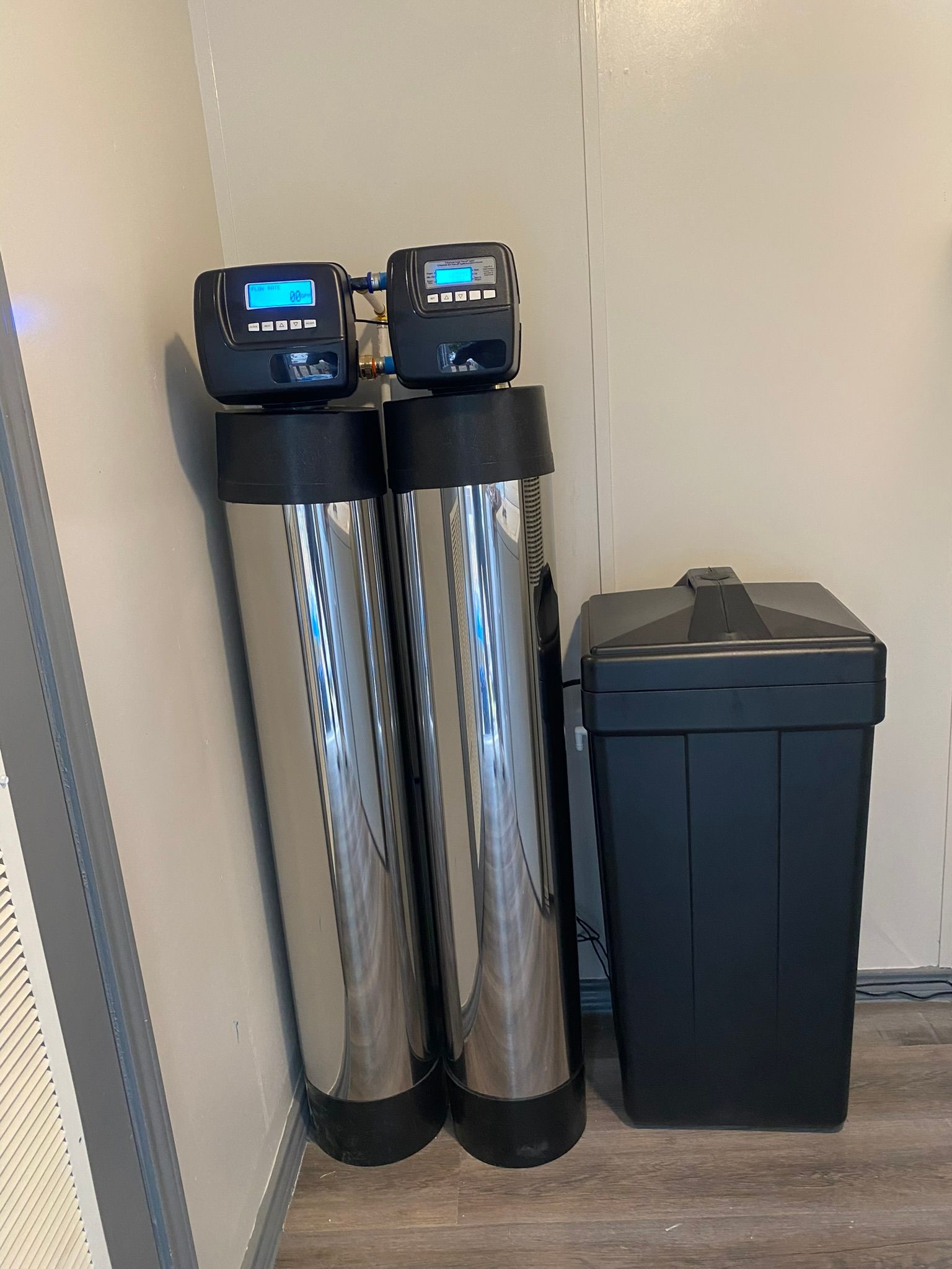 Water softener system in a basement. A tall blue tank stands next to a white salt tank. Pipes and control panel are visible.