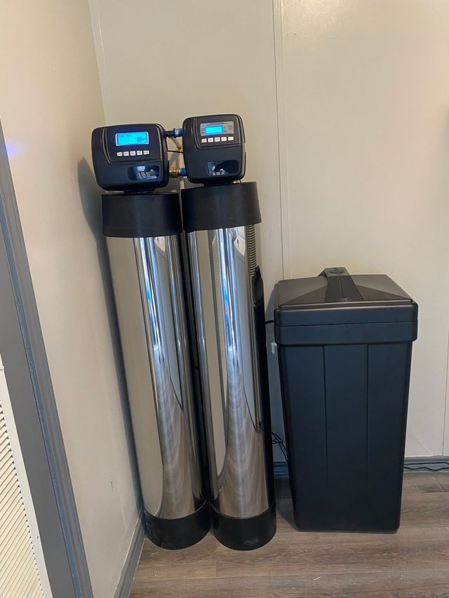 Water softener system in a basement. A tall blue tank stands next to a white salt tank. Pipes and control panel are visible.