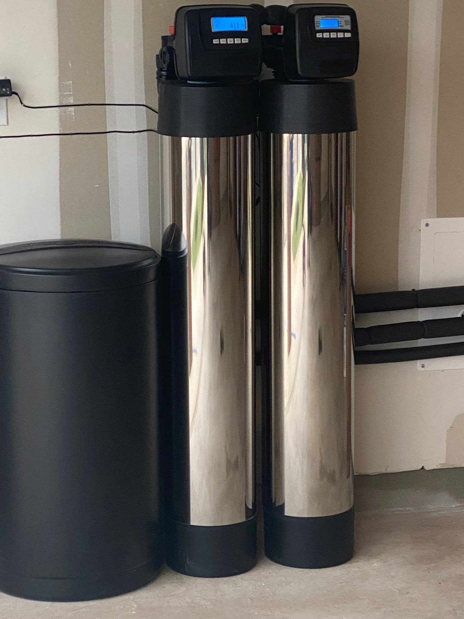 Water filtration system: two tall chrome tanks with digital displays, black brine tank, against a light wall.