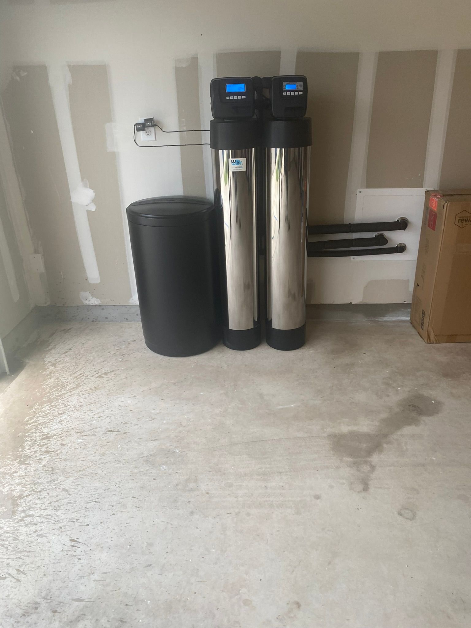 Water softener system in a garage. Two tall silver tanks, a black salt tank, and exposed pipes.