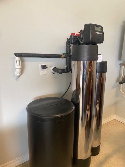 A water filtration system with two silver tanks, a black control head, and a black salt storage tank against a beige wall.