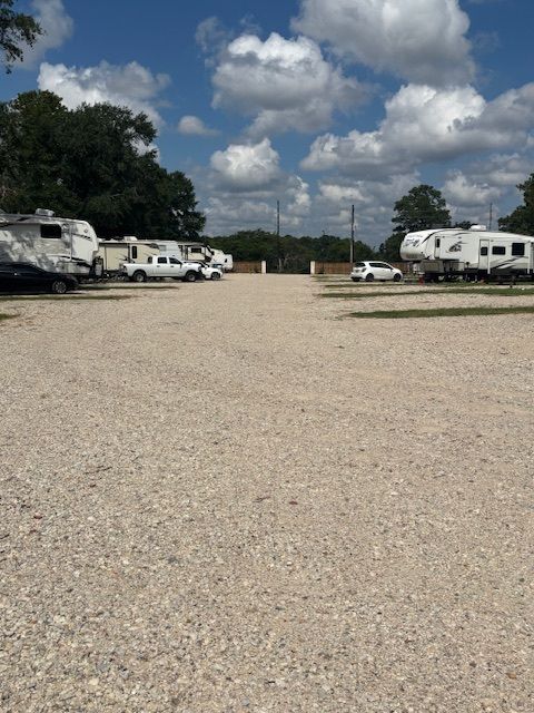 RV storage lot with gravel, RVs, white trucks, gate, blue sky with clouds.