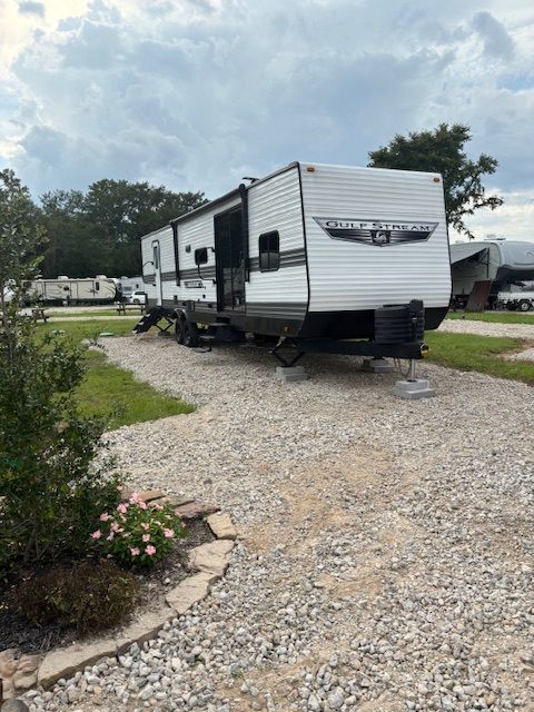 RV parked on gravel at a campsite, with slide-out extended. Cloudy sky overhead.