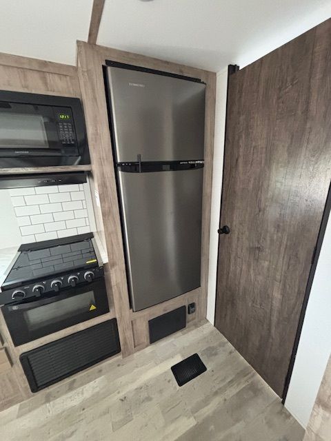 RV interior: Stainless steel refrigerator next to a wood-grain door, beside a stove and microwave.