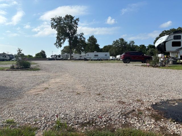 Gravel RV parking area with several RVs parked. A red SUV is parked on the gravel, under a blue sky.
