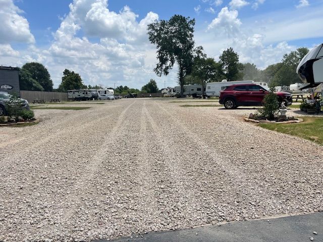 Gravel RV campground with RVs parked, red SUV, trees, and blue sky.