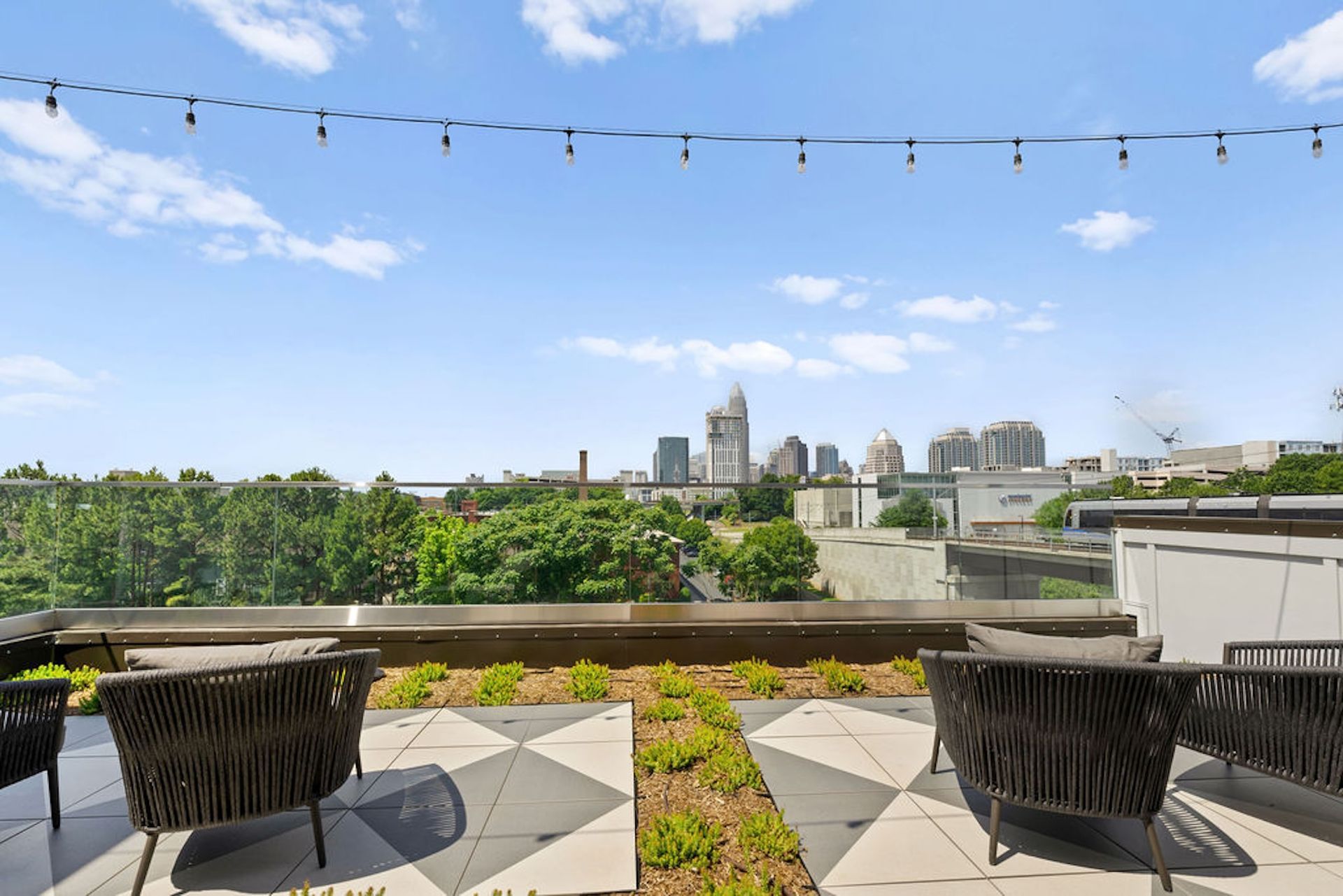 Rooftop patio with city skyline view, featuring chairs, string lights, and a cloudy blue sky.