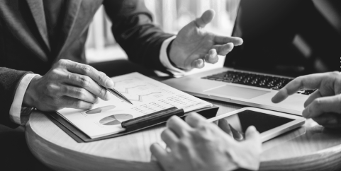 Black and white image of 2 people discussing with a paper on the table with a lot of charts.