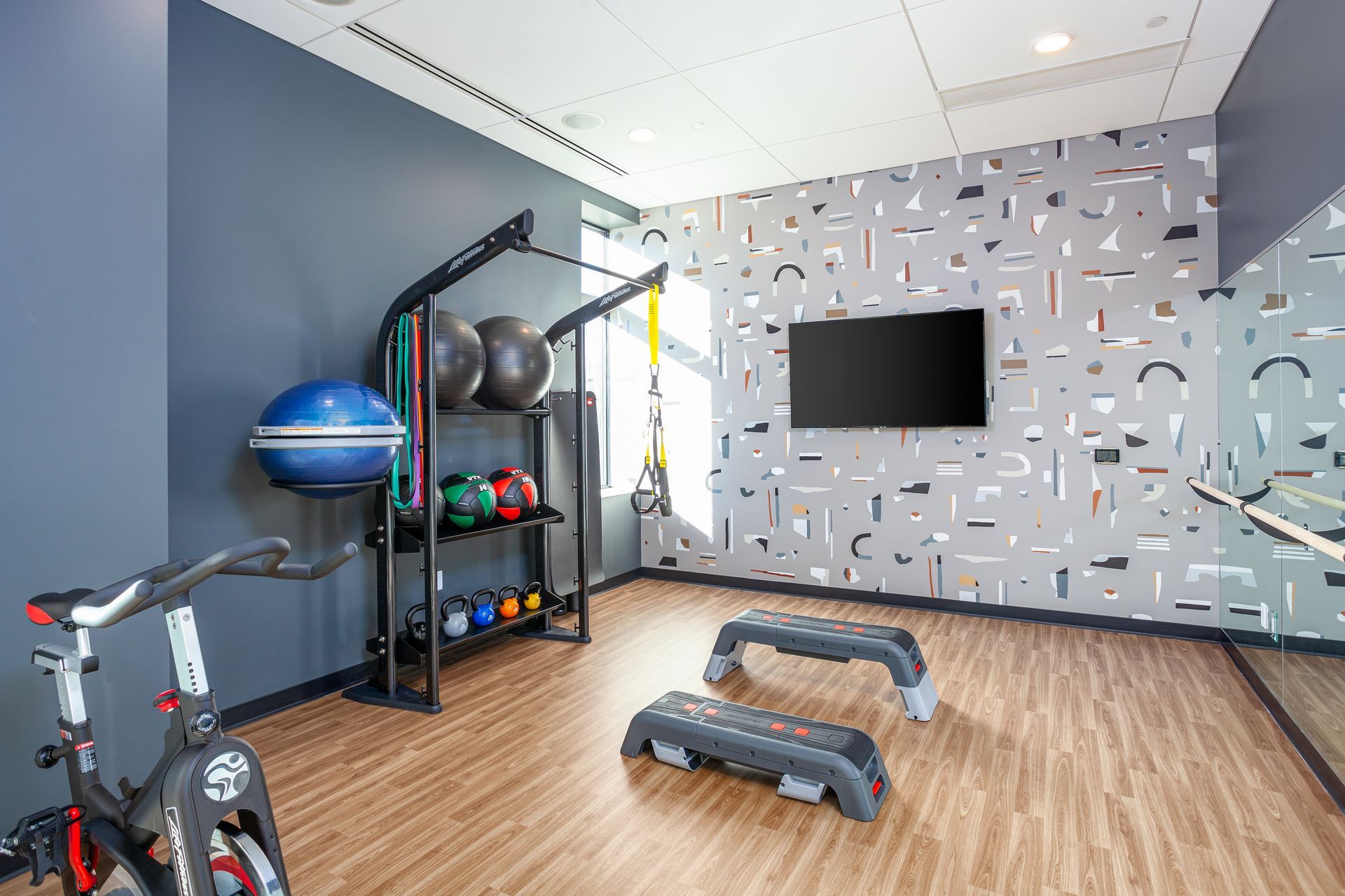 Gym with exercise equipment: stationary bike, weights, steps, TV, patterned accent wall, and mirror.
