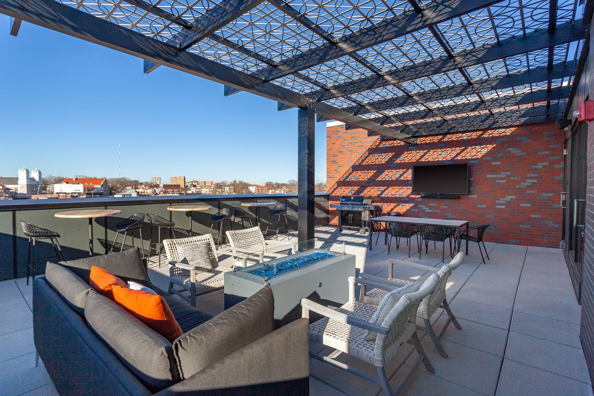 Rooftop patio with seating, fire pit, grill, and TV under a patterned canopy, overlooking a cityscape on a sunny day.