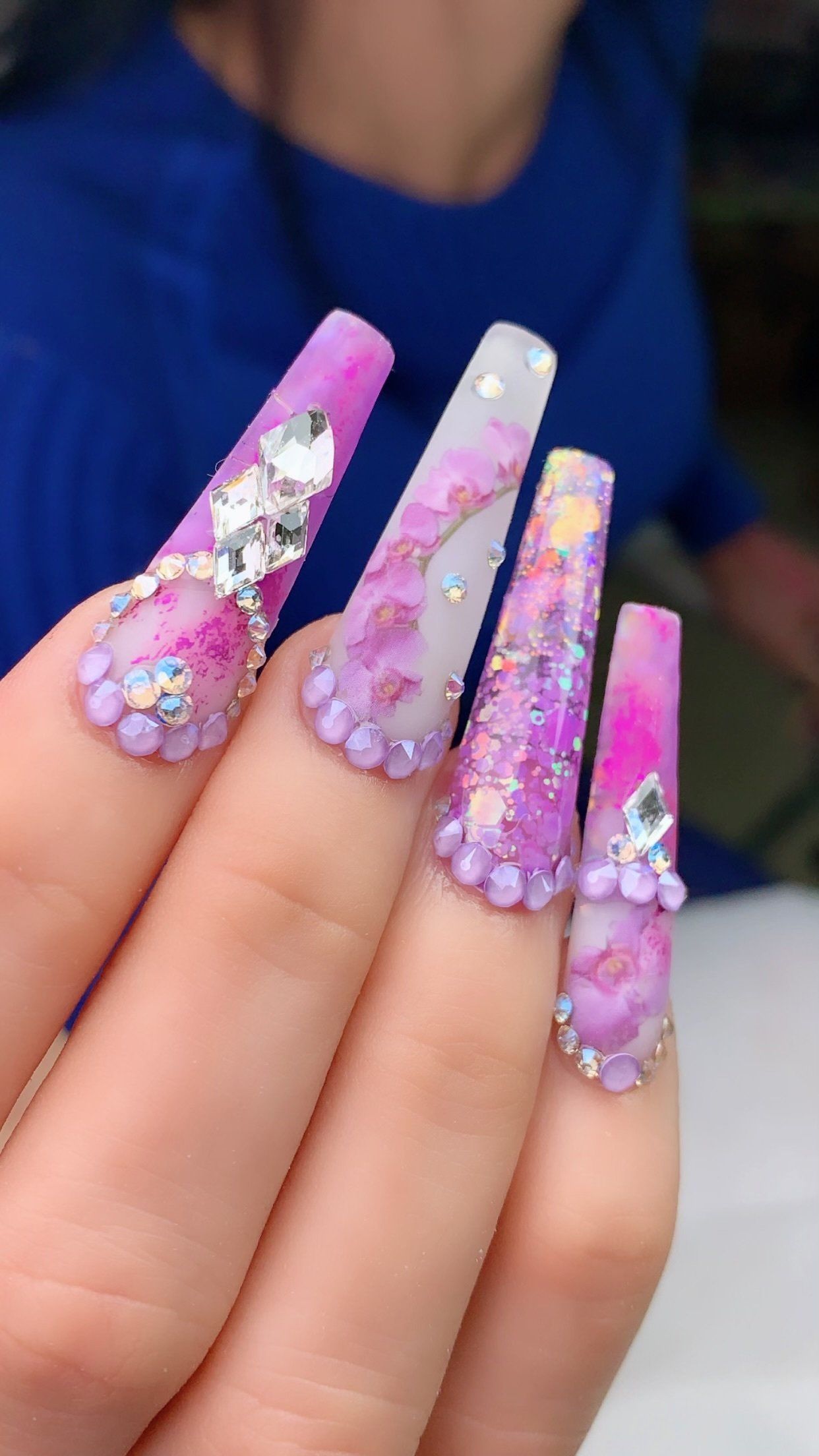 A person is holding a pair of long purple nails with rhinestones.